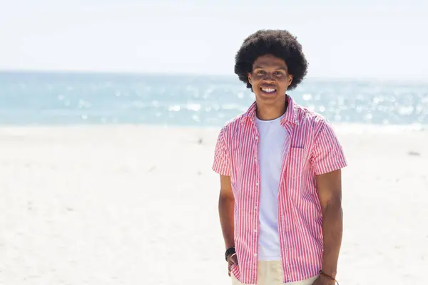 A young African American man smiles at the beach, with copy space unaltered. He enjoys a sunny day outdoors by the sea.