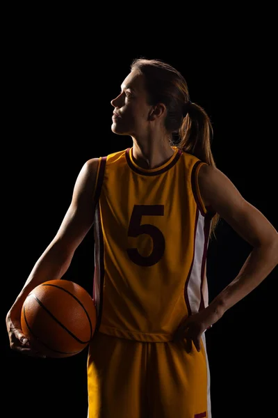 Young Caucasian female basketball player poses confidently in a basketball uniform on a black background. She holds a basketball, symbolizing determination and athleticism.