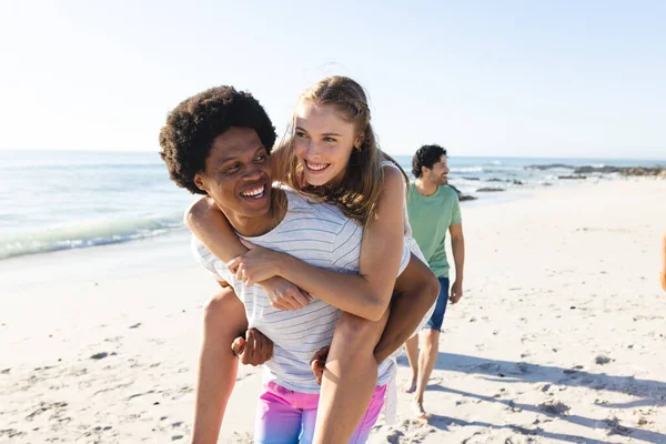 Young African American man gives a piggyback ride to a Caucasian woman on the beach. Their laughter and playful energy enhance the carefree outdoor setting.