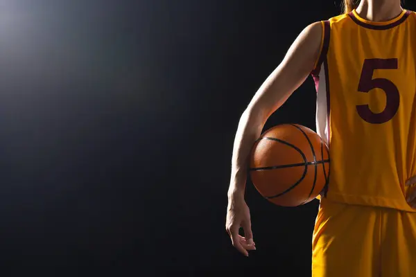Athlete in basketball uniform holds a ball on a black background, with copy space. Captured in a studio setting, the focus is on sports and athleticism.