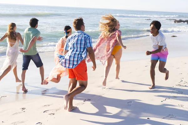Diverse group of friends enjoy a beach outing. They are running along the shoreline, embodying a carefree summer vibe.