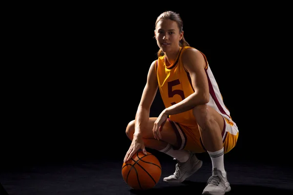 Young Caucasian female basketball player poses confidently in basketball attire, with copy space on a black background. Captured in a studio setting, she exudes athletic prowess and focus.