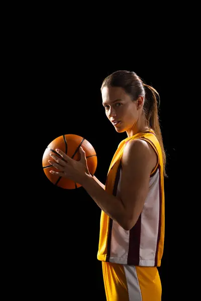 Young Caucasian female basketball player poses confidently in basketball attire, with copy space on a black background. She\'s ready for a game, showcasing skill and focus in a dark setting.