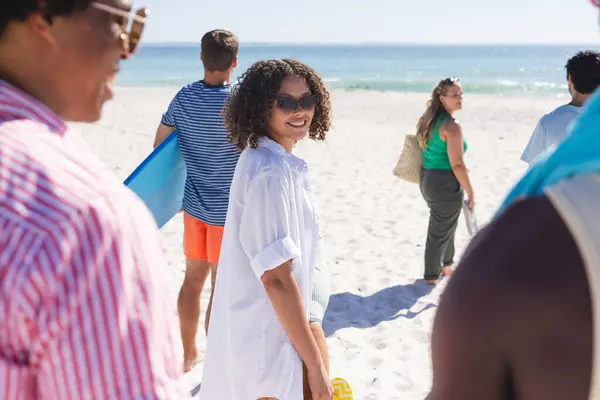 A diverse group of friends enjoys a sunny day at the beach. Smiling young Biracial woman stands out in a casual white shirt, surrounded by diverse companions.