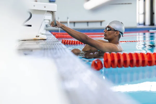 Young biracial male athlete swimmer prepares for a swim at an indoor pool. His focus on performance highlights the competitive nature of swimming.