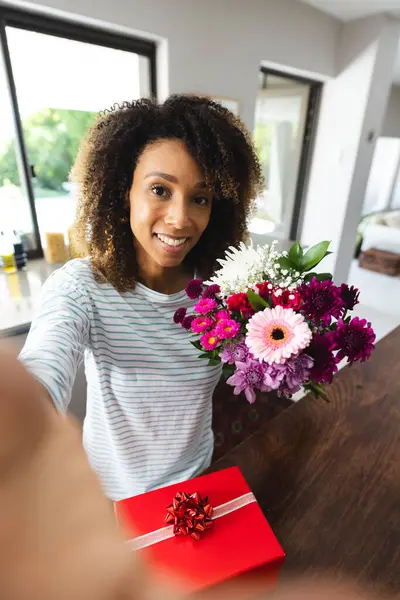 Biracial woman takes a selfie at home. She\'s smiling with a bouquet and a gift, capturing a joyful moment.
