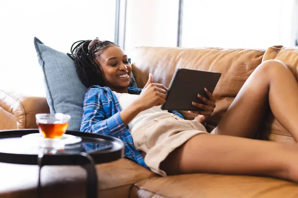Young African American woman enjoys a tablet on a cozy couch, tea on the table. Relaxed home setting with natural light, she exhibits a leisurely and content mood.