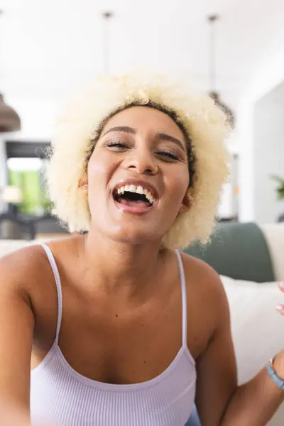 Young biracial woman with blonde hair laughs joyfully on video call, wearing a white tank top. Captured indoors, her expression exudes happiness and relaxation at home.