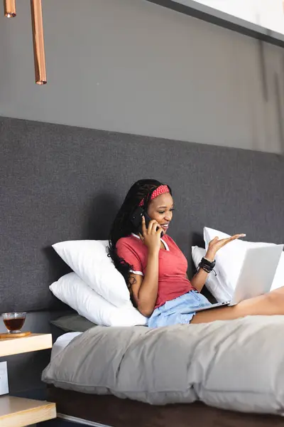 Young African American woman chats on the phone while using a laptop in bed with copy space. She\'s wearing a red headband, casual clothes, and appears relaxed in a cozy home setting.