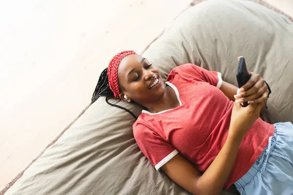 一位年轻的非洲裔美国妇女一边笑一边用她的智能手机 她躺在米黄色的垫子上 身穿红色上衣 头戴斜纹棉布 在家里散发出轻松的光芒 — 图库照片