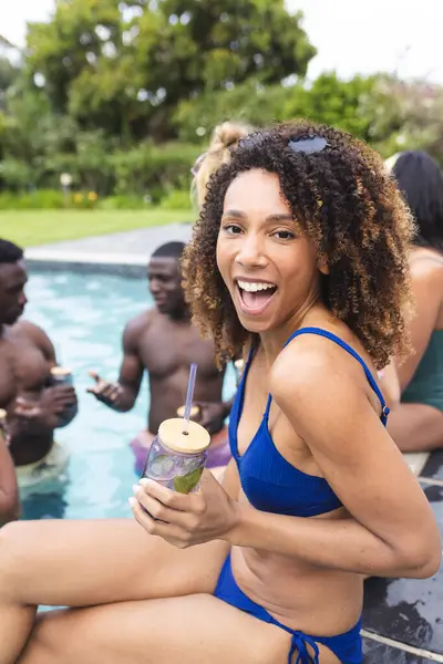 Biracial woman in a blue swimsuit enjoys a pool party, holding a drink with a smile. Her curly brown hair and joyous expression capture the lively outdoor summer vibe.