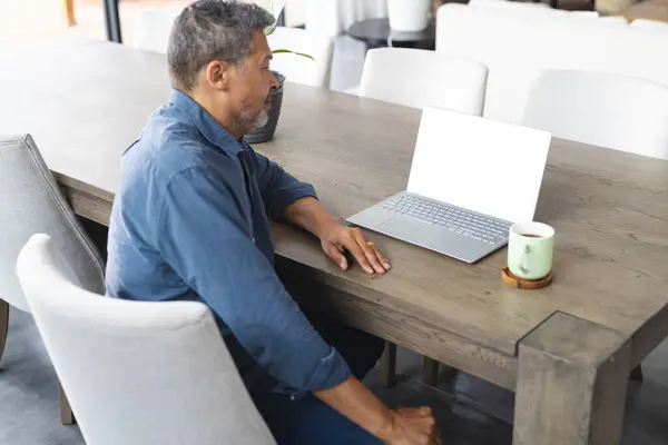 stock image Biracial senior man works on a laptop at a modern dining table with copy space. He appears focused, with salt-and-pepper hair, in a casual blue shirt.