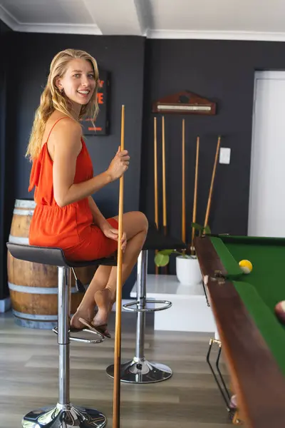 身穿橙色衣服的年轻白人女人喜欢玩撞球游戏 手里拿着一根带有复制空间的球杆 她金发碧眼的发型和随意的姿势暗示了游戏室里一种轻松的气氛 — 图库照片