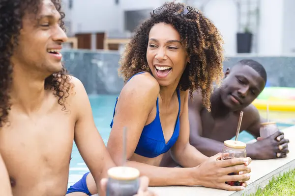 Biracial woman and men enjoy a poolside gathering. She laughs in a blue swimsuit, while the young men relax with drinks, sharing a joyful moment.