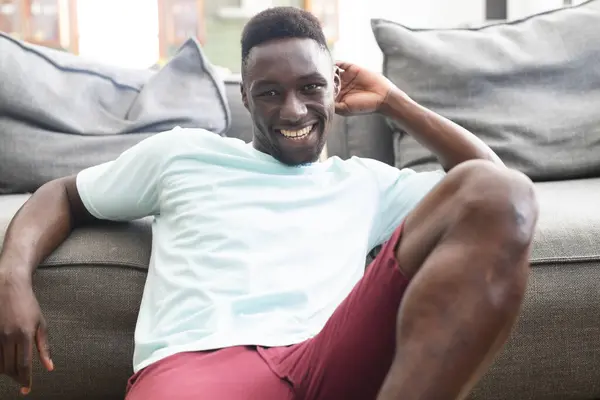 年轻的非洲裔美国人躺在沙发上 面带微笑 他穿着休闲的浅蓝色T恤和红色短裤 在家里散发出放松的感觉 图库照片