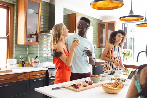 Young Caucasian woman in an orange dress laughs with a young African American man holding a drink. Biracial woman, busy preparing food, glances over at them in a modern kitchen setting.
