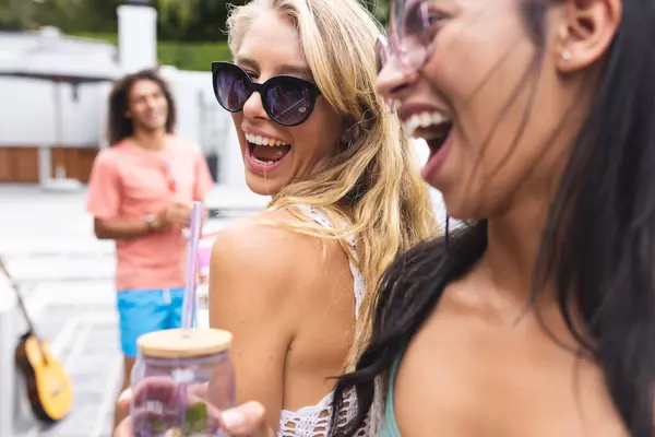 Laughing young Caucasian woman with blonde hair and sunglasses is with a biracial woman. They enjoy a sunny day outdoors, with a young biracial man smiling in the background.