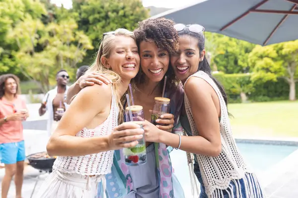 三个不同的女性朋友在户外泳池派对上拥抱和微笑 不同的朋友在一起享受一个阳光灿烂的日子 背后是一位年轻的非裔美国人 免版税图库照片