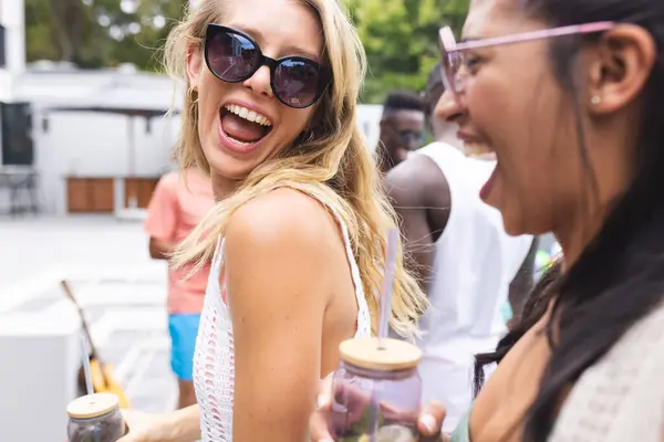戴着太阳镜的年轻白人妇女和一位年轻的非洲裔美国妇女在户外分享欢笑 他们在一起享受着一个阳光灿烂的日子 散发着幸福和友情 免版税图库照片