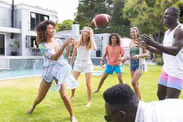 多样化的朋友圈在阳光灿烂的后院欣赏一场足球比赛 年轻的白人女子抛出足球时 空气中充满了笑声 年轻的非洲裔美国人准备接住足球 图库照片