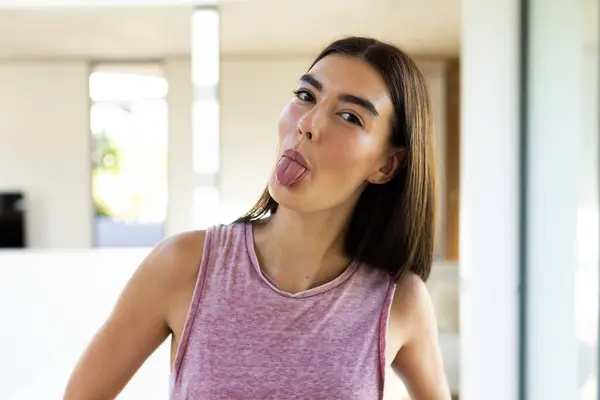 A young Caucasian brunette woman sticks her tongue out playfully at home. She has long brown hair, a sleeveless top, and exhibits a carefree attitude, unaltered.