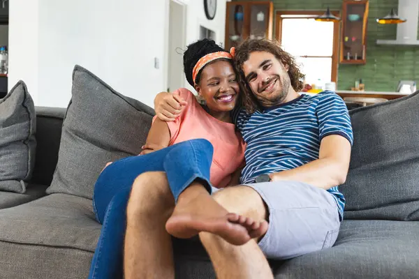 一对多样化的夫妇在家里的沙发上分享着一个舒适的时刻 她是个非洲裔美国人 面带笑容 头带和橙色上衣 而他的卷发和条纹衬衫 — 图库照片