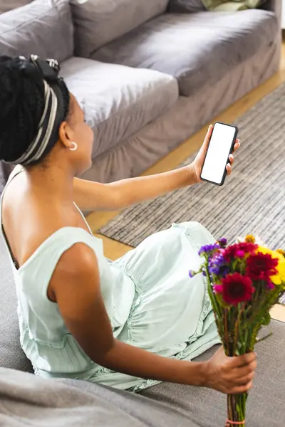 stock image African American woman holding smartphone, showing screen on a video call date with copy space. She has dark hair, wearing a light green dress, and holding colorful flowers, unaltered.