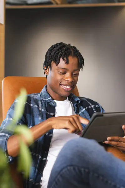 非洲裔美国人坐在一个现代的商业办公室里 用一种带着微笑的平板电脑 他穿着休闲装 留着短短的扭曲的头发 年轻的样子 没有变 免版税图库图片