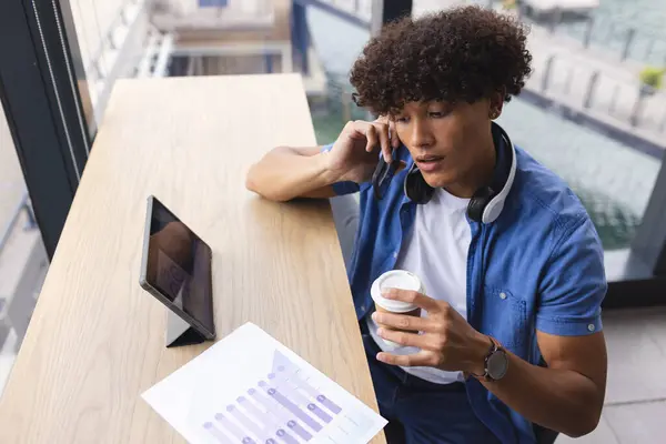 一个留着卷曲棕色头发的年轻的比尔卡利亚男子正在一家现代商务办公室打电话 他穿着一件蓝色衬衫 拿着一杯咖啡 还有一块刻有Vr图形的平板电脑 没有改动 图库图片