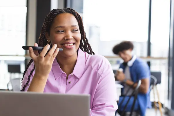 在一个现代化的商业办公室里 一个会用智能手机说话 会在笔记本电脑上工作的年轻的比利西亚女人 她编了辫子 穿了件粉色的衬衫 背景上站着一个年轻的黑人男子 免版税图库照片