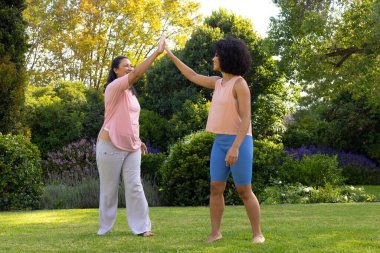 Biracial mother and daughter are high-fiving in garden at home. Both wearing casual clothes, young woman has curly hair, the mature woman straight, unaltered clipart