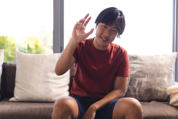 亚洲少年坐在家里的沙发上 在视频通话中做手势 穿着红色T恤 黑头发 看着相机 图库图片