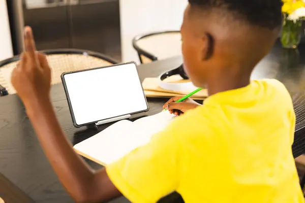 非裔美国男孩专注于在家写作与复制空间 平板与空白屏幕附近 他有一头黑色的短发 穿着黄色的衬衫 图库照片
