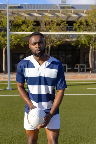 African American young male athlete holding rugby ball on field outdoors, looking serious. He has short black hair, beard, and is wearing sporty clothes, unaltered.