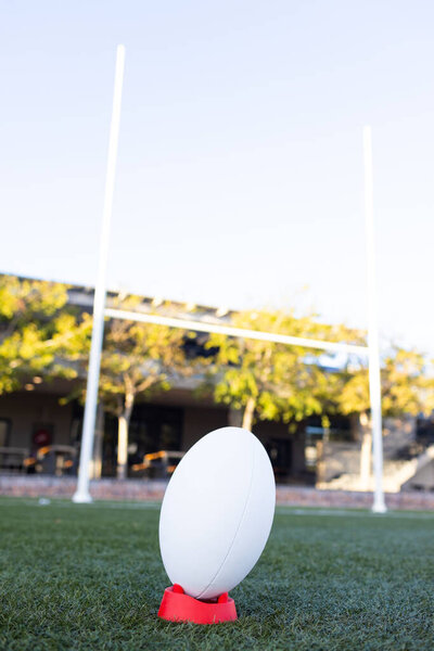 Rugby ball standing on red kicking tee on field outdoors, field goal posts in background, copy space. Sunlight bathing field in warm light, clear day for sports, unaltered. — Stock Image Rugby Ball Standing Red Kicking Tee Field Outdoors Field Goal Royalty Free Stock Images