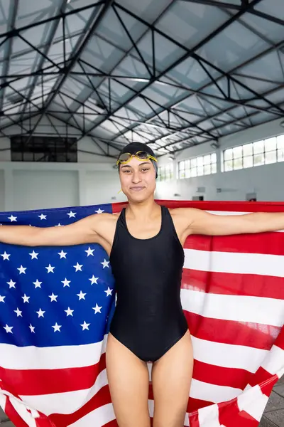 年轻的年轻女子游泳运动员站在室内 裹着美国国旗 对着镜头微笑 她身材很健康 穿着黑色泳衣 帽子和护目镜 没有改变 免版税图库图片