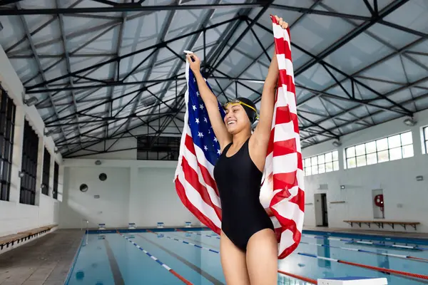 年轻的年轻女子游泳运动员举着美国国旗站在空旷的室内 复制空间 她的皮肤是褐色的 头发是黑色的 身穿泳衣 头戴一顶泳帽 没有变 免版税图库照片
