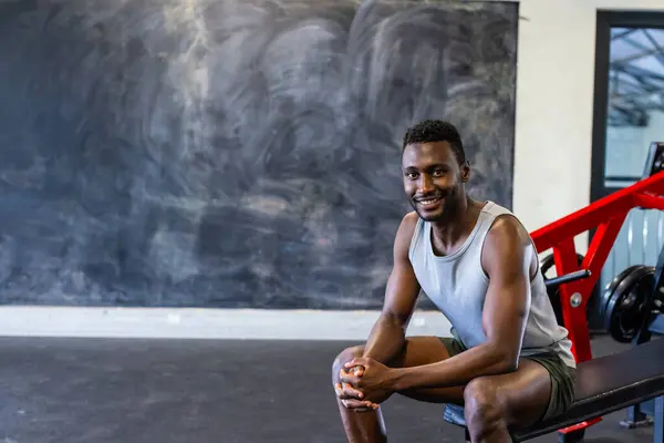 非裔美国青年男子运动员坐在体育馆的长椅上 微笑着 复制着空间 他身穿背心和短裤 看上去强壮而健康 没有改变 图库图片