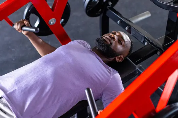 一位年轻的非洲裔美国男子运动员 躺在体育馆的长椅上举重 他有一头黑色的短发 留着胡子 穿着一件紫色的T恤 图库图片