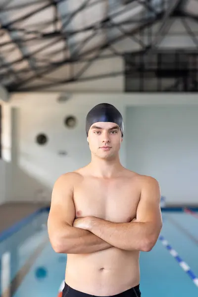 白人年轻男子游泳运动员站在室内游泳池边 双臂交叉 看着相机 复制空间 他有一头棕色的短发 皮肤白皙 戴着一顶黑色的游泳帽 一点也没变 免版税图库图片