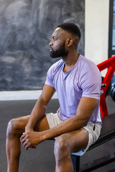 非裔美国青年男子运动员坐在体育馆的长椅上 复制空间 他有短短的黑头发 留着胡子 穿着紫色衬衫和短裤 图库照片