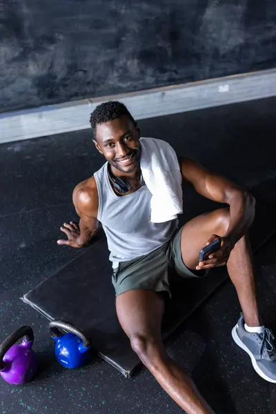 非洲裔美国男性运动员和健身模特儿在健身房 手持智能手机 运动后休息 他有一头黑色的短发 身穿背心上衣和短裤 面带微笑 没有变 免版税图库图片