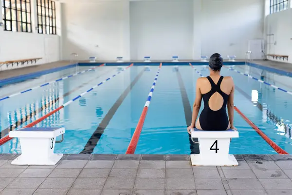 幼小的年轻女子游泳运动员坐在室内游泳池边 看着水 抄袭空间 她有一头乌黑的头发 穿着黑色的泳衣 随时准备训练 毫发无损 图库图片
