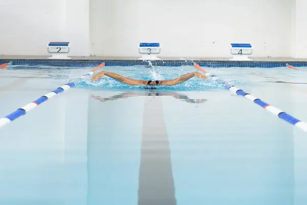 Biracial young female swimmer reaching end of the swimming pool lane, copy space. Water splashing around as she completes lap indoors, unaltered