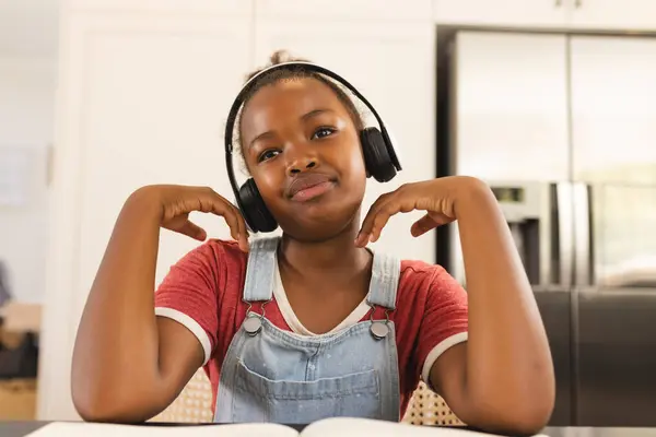 非洲裔美国女孩戴着耳机 在家里欣赏音乐 手肘搁在红底桌子上 穿着红衫和斜纹棉布工作服 看上去心满意足 图库照片
