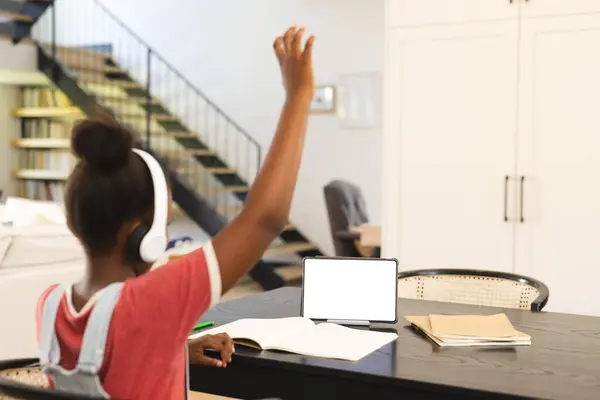 ヘッドフォンのアフリカ系アメリカ人の少女は タブレットベースのオンラインレッスン コピースペースで手を挙げます 居心地の良いタッチ ラップトップ テーブル上の本を飾る木製のフレーム ロイヤリティフリーのストック画像