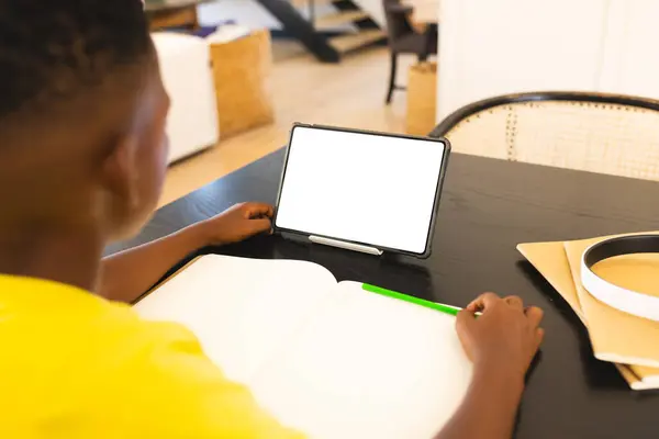 自宅では アフリカ系アメリカ人の少年が緑色のペンを持ち タブレットを見て スペースをコピーします 赤いシャツを着て ホワイトバックグラウンドのオープンノートに書き込む準備ができています ストック写真