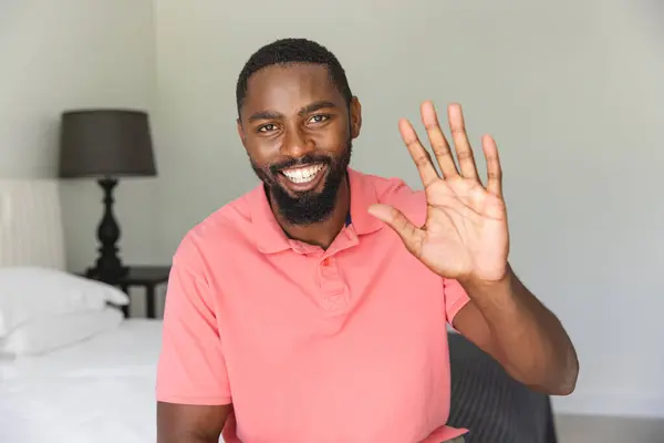 非洲裔美国人在一个视频通话中挥手 坐在家里的卧室里 他留着胡子 穿着一件粉红色的衬衫 热情地笑着 没有变 免版税图库图片