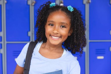 Biracial girl smiles in a school setting. Her cheerful expression brightens the hallway lined with blue lockers. clipart