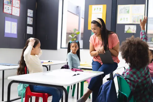 stock image In school, young biracial female teacher engaging with diverse students in classroom. She has dark hair, wearing casual clothes, holding tablet, learning, education, unaltered